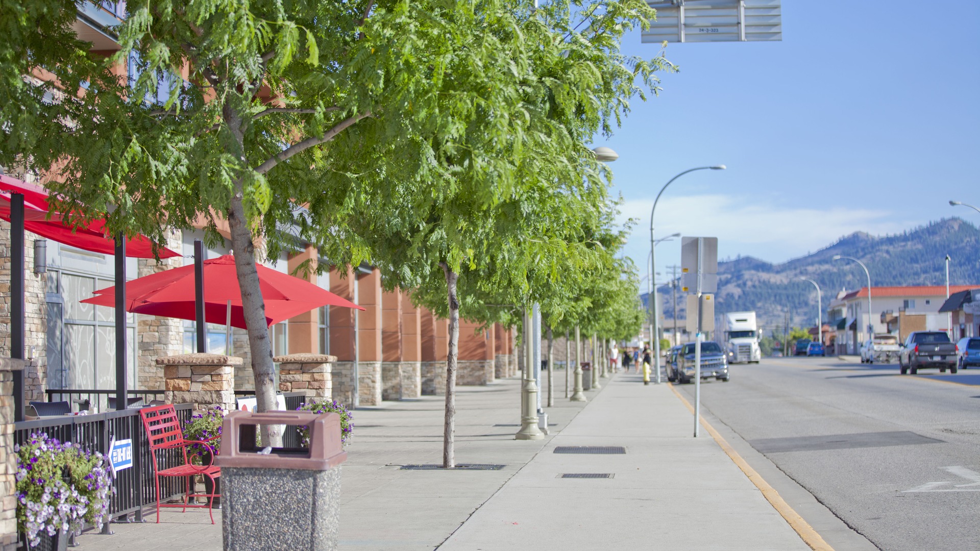 Tree-lined downtown sidewalk and street in Osoyoos with low-rise buildings and hills beyond