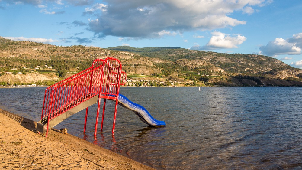 Red and blue beach slide at Skaha Lake with water and Okanagan hills beyond